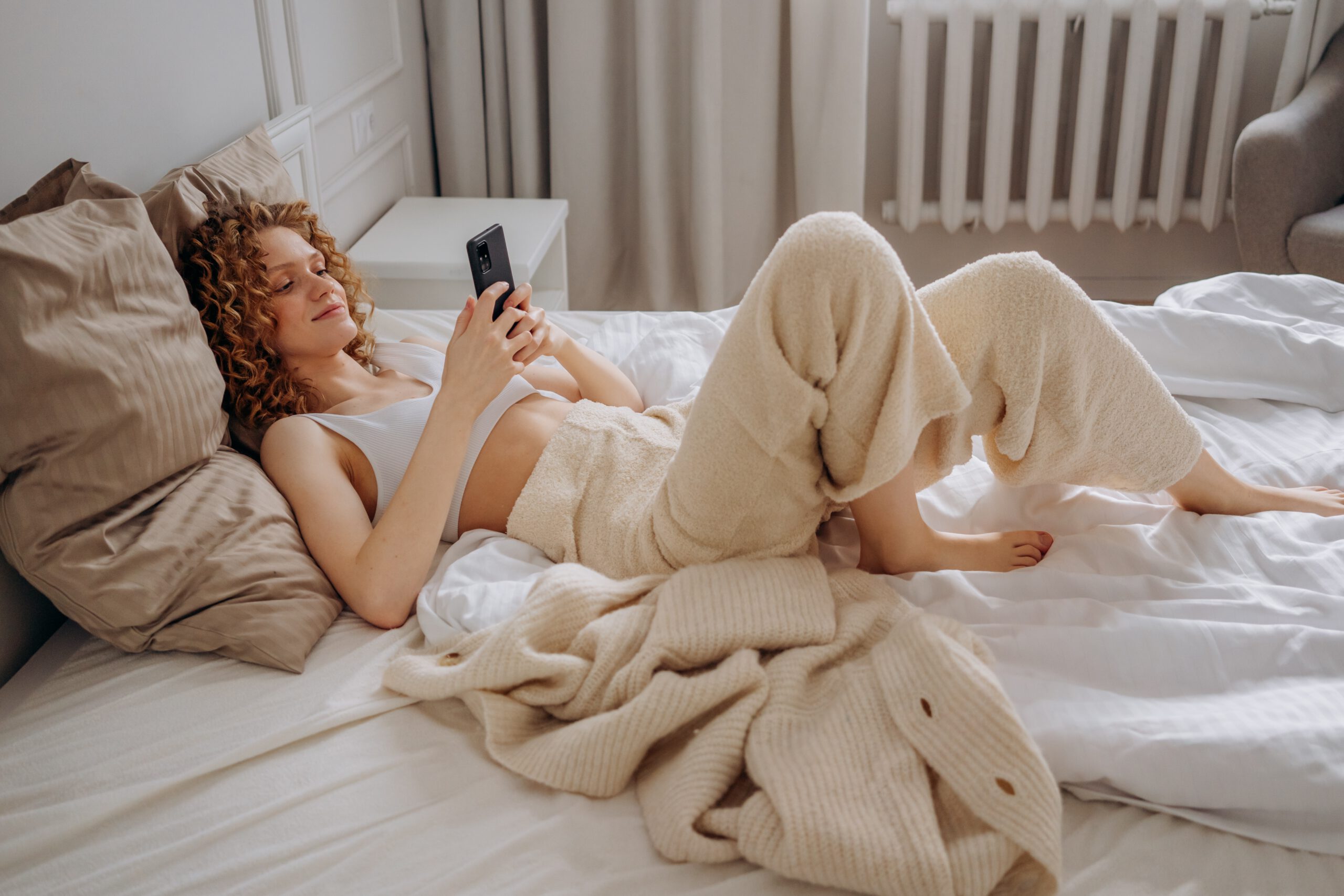 Kobieta leżąca na łóżku w białym topie i luźnych spodniach z telefonem w ręce
